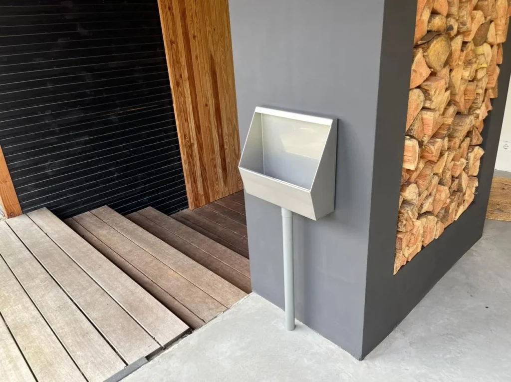 RVS tuinurinoir gemonteerd op grijze wand bij houtopslag onder overkapping