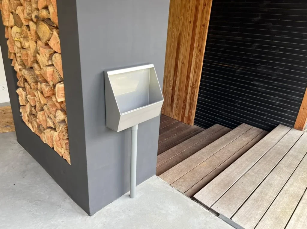 RVS tuinurinoir tegen donkergrijze wand naast gestapelde houtblokken en houten lattengevel