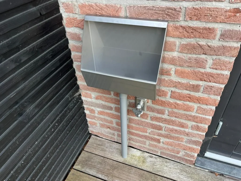 RVS tuinurinoir tegen rode bakstenen muur op houten terras met tapkraan eronder