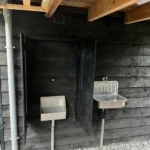Klein RVS urinoir met grote houten privacy-panelen en RVS wasbak-fontein in een buitenkeuken
